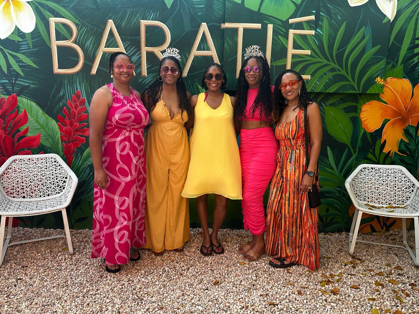 Five travelers in bright summer outfits and birthday tiaras in front of a tropical mural